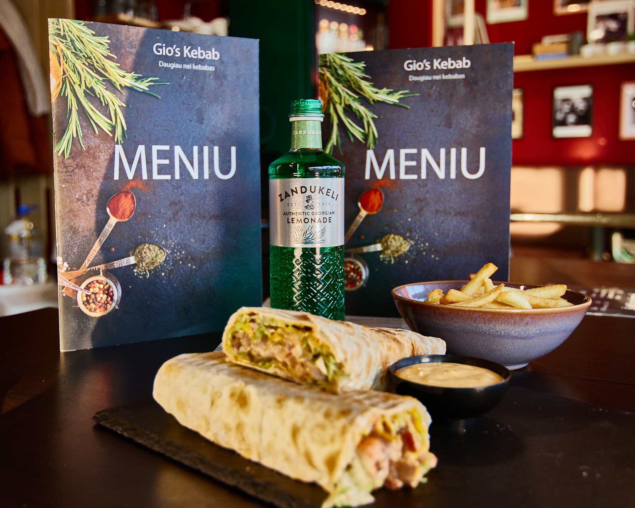 Maisto serviruotės reklaminis kadras Ant stalo pateiktas kebabas, bulvytės, padažas ir gėrimas restorano aplinkoje